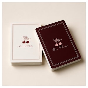May include: Two stacks of rectangular cards. The left stack is cream-colored with a dark red cherry illustration, a double-line border, and the text Hannah Miller. The right stack is dark red with a light pink cherry illustration and Ms. Thomas.