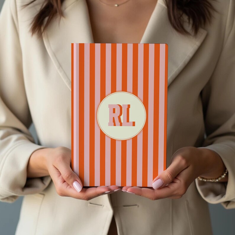 Orange and pink striped personalized notebook held by a woman, featuring custom initials on the front cover. Modern monogrammed journal with perforated lined pages for writing, journaling, or business notes. Elegant hardcover notebook for students, professionals, or creatives. Aesthetic orange stripe stationery perfect for office use, school, or gift-giving. Custom name notebook ideal for thoughtful personalized gifts or stylish daily planning.
