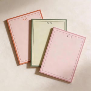May include: Three rectangular notepads in shades of pink and green, each with a contrasting border and initials in the upper corner. The notepad colors are pink, light green, and pink. The initials are N.O., W.S., and C.G.