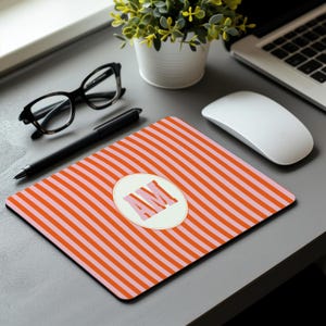 May include: A rectangular mousepad with a striped pattern in alternating shades of orange and pink. The mousepad features a white oval with the letters "AM" in pink. Other items on the desk include glasses, a pen, and a computer mouse.