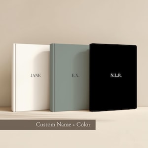May include: Three hardback books in white, grey-green, and black. Each book has a name or initials printed in a simple font. The text "Custom Name + Color" is at the bottom of the image.