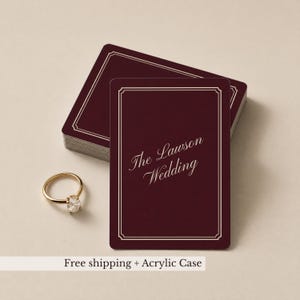 May include: A stack of rectangular deep burgundy cards with a thin white border and the text The Lawson Wedding in a white script font, accompanied by a gold ring featuring a single clear stone set on a thin, polished gold band.
