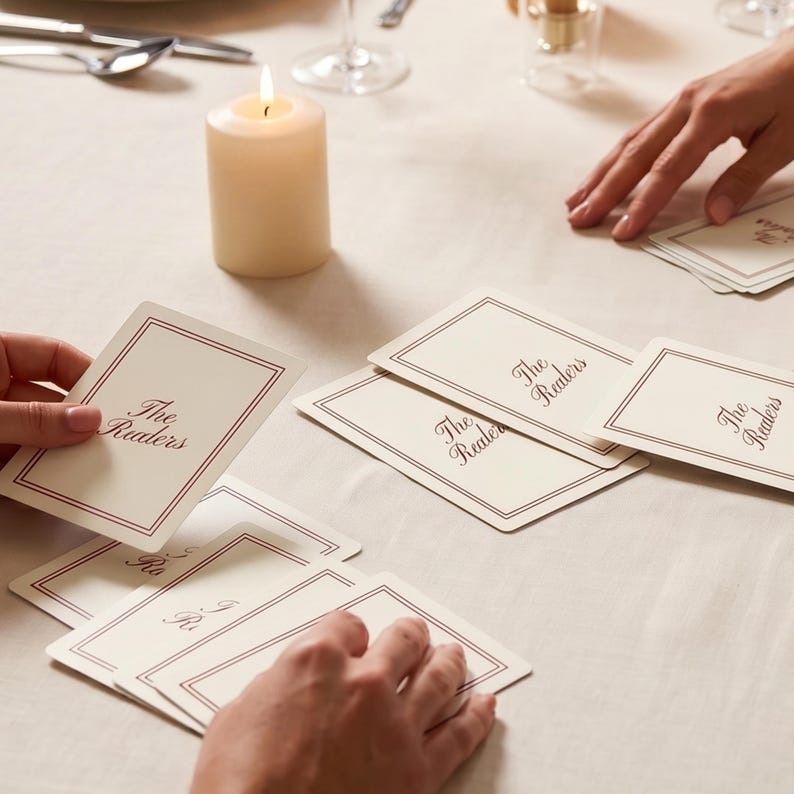 May include: A rectangular deck of off-white playing cards with a dark red double-line border. The text "Tina's 28th Birthday" is printed in dark red script in the center of the top card. A white ranunculus flower is positioned to the left.