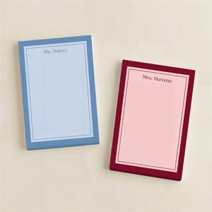 May include: Ivory stationery with a light blue border and the initials "E.F." is displayed on a marble surface. A gold pen rests on a light pink envelope. A cup of coffee and a vase with flowers are also present.
