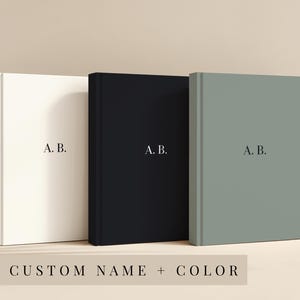 Op de afbeelding: Drie hardcover journals in crème, zwart en saliegroen, elk met de initialen "A. B." gedrukt in een minimalistische stijl. De tekst "CUSTOM NAME + COLOR" staat onderaan. De journals worden rechtop naast elkaar weergegeven.