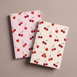 Op de afbeelding: Twee notitieboeken met harde kaft en een herhalend kersenpatroon. Het ene notitieboek heeft een lichtroze kaft met rode kersen, terwijl het andere een crèmekleurige kaft met rode kersen heeft. Beide notitieboeken hebben een rechthoekige vorm.