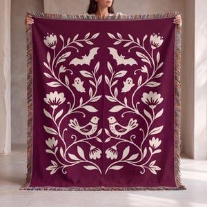 May include: A deep burgundy woven throw blanket featuring a symmetrical cream-colored pattern of floral vines, birds, bats, and ghosts. The rectangular blanket has a fringed edge along the top and bottom, showcasing a multi-colored thread detail.