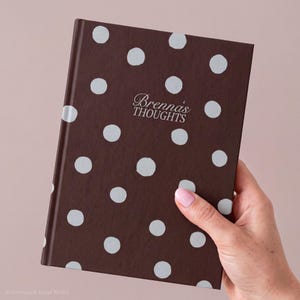 May include: A dark brown hardcover journal featuring a repeating pattern of light gray polka dots. The front cover displays the text "Brenna's THOUGHTS" in a white serif font centered among the polka dot pattern.