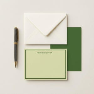May include: Stationery set featuring a pale green notecard with a dark green border and the name "JOHN BRIGHTON" printed at the top. Includes a white envelope, a dark green card, and a black and gold pen.