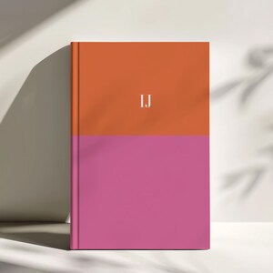 A personalized colorblock notebook with a bold orange top half and vibrant pink lower half stands upright in soft studio lighting with leaf shadows cast on the background. White monogram initials appear centered on the cover. This modern bright design is ideal for creative planners students writers and gift giving. The clean minimalist hardcover layout fits premium stationery collections and high quality desk accessories with vibrant trendy colors.