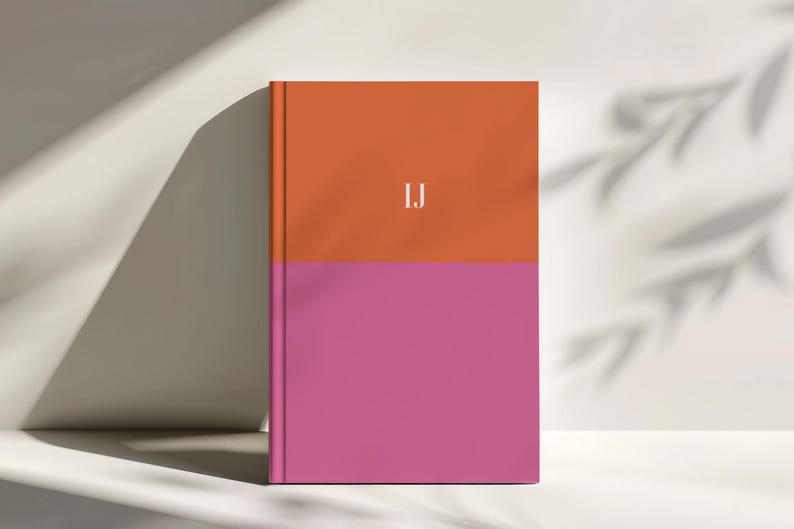 A personalized colorblock notebook with a bold orange top half and vibrant pink lower half stands upright in soft studio lighting with leaf shadows cast on the background. White monogram initials appear centered on the cover. This modern bright design is ideal for creative planners students writers and gift giving. The clean minimalist hardcover layout fits premium stationery collections and high quality desk accessories with vibrant trendy colors.