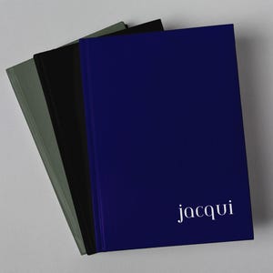 May include: Three hardback journals stacked together. The top journal is deep blue with the name jacqui printed in white lowercase letters. The middle journal is solid black, and the bottom journal is a muted olive green colour.