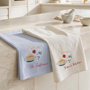 May include: Two rectangular kitchen towels, one light blue and one off-white, each featuring a printed graphic of a plate of spaghetti, a tomato, and the text la dolce vita. The blue towel reads The Jeffersons and the white towel reads Tyra's Kitchen.