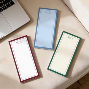 May include: White stationery with a navy blue border and the initials "J.K." in the upper center. The stationery is on a marble surface, with office supplies such as pencils and binder clips in the background.
