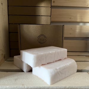 May include: Three rectangular bars of white soap are stacked on a wooden surface. A brown cardboard box with a circular logo is behind the soap. Additional boxes are stacked in the background, suggesting a product display.