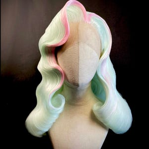 Puede incluir: Peluca de color pastel con base verde claro y mechas rosas, peinada con rizos largos y ondulados. La peluca se exhibe sobre una cabeza de maniqu&iacute; sobre un fondo oscuro. La peluca tiene una parte delantera de encaje y est&aacute; etiquetada "Stephane SCOTTO DI CESARE".