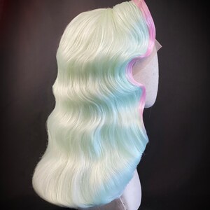 Puede incluir: Una peluca de colores pastel con cabello ondulado hasta los hombros. La peluca presenta una mezcla de verde claro, azul y blanco, con un borde rosa. La peluca se muestra sobre una cabeza de maniqu&iacute; sobre un fondo oscuro. El texto "By Stephane SC0TTO DI CESARE" est&aacute; en la parte inferior.