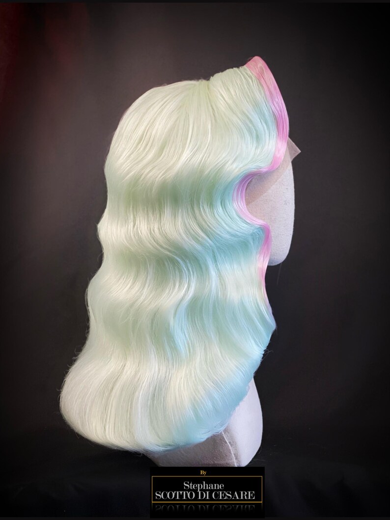 Puede incluir: Una peluca de colores pastel con cabello ondulado hasta los hombros. La peluca presenta una mezcla de verde claro, azul y blanco, con un borde rosa. La peluca se muestra sobre una cabeza de maniqu&iacute; sobre un fondo oscuro. El texto "By Stephane SC0TTO DI CESARE" est&aacute; en la parte inferior.