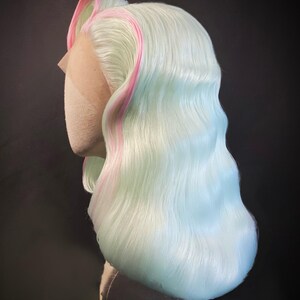 Puede incluir: Una peluca de colores pastel con una base azul claro y verde menta, acentuada con reflejos rosas. La peluca est&aacute; peinada con ondas suaves y una raya a un lado. El encaje frontal es visible y la peluca se muestra en una cabeza de maniqu&iacute;. La etiqueta dice "By Stephane SCOTTO DI CESARE".