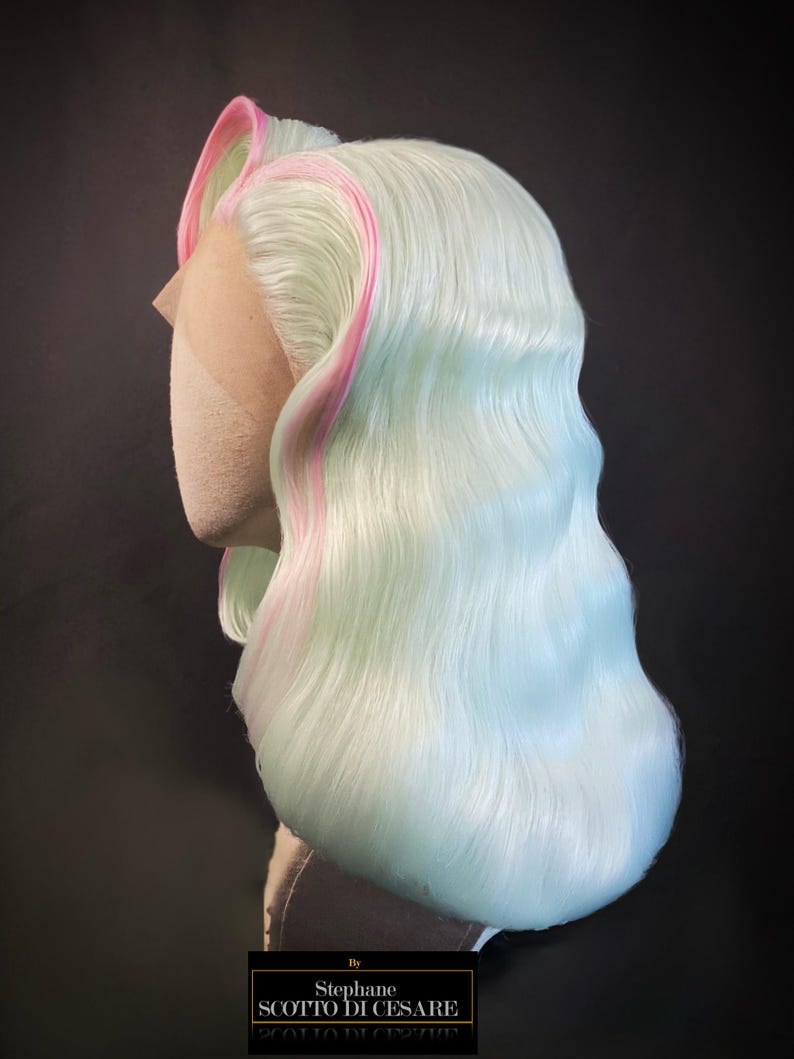 Puede incluir: Una peluca de colores pastel con una base azul claro y verde menta, acentuada con reflejos rosas. La peluca est&aacute; peinada con ondas suaves y una raya a un lado. El encaje frontal es visible y la peluca se muestra en una cabeza de maniqu&iacute;. La etiqueta dice "By Stephane SCOTTO DI CESARE".