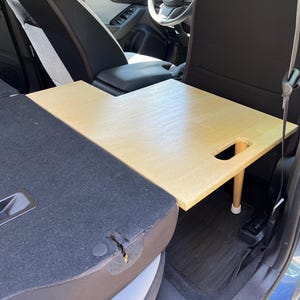 Puede incluir: Una mesa de coche de madera de color claro con un asa recortada y una sola pata. La mesa está colocada en el asiento trasero de un coche, con el asiento y el interior visibles. La mesa mide aproximadamente 61 cm de largo y 41 cm de ancho.