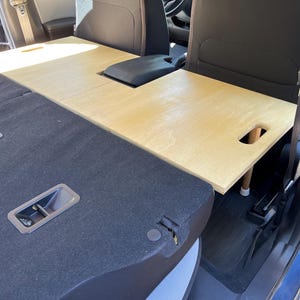 Puede incluir: Una mesa de asiento de coche de madera de color claro con un recorte para la consola central. La mesa está soportada por pequeñas patas y está diseñada para encajar entre los asientos delanteros y traseros de un vehículo.