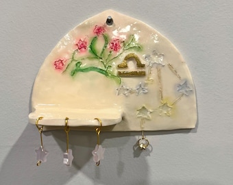 Può includere: Una mensola da parete in ceramica color crema a forma di semicerchio con un motivo floreale rosa dipinto, un simbolo dorato del segno zodiacale Bilancia e una costellazione. Tre piccoli ganci dorati sostengono ciondoli a stella.