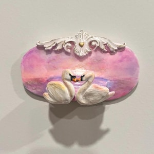 Può includere: Decorazione murale ovale con due cigni di fronte a un tramonto rosa e viola. La parte superiore della decorazione presenta un design ornato bianco e oro. Scena romantica.