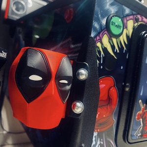 May include: A red and black Deadpool mask attached to a pinball machine. The pinball machine has a colorful design with a "Start" button and Deadpool graphics. The mask is mounted with silver screws.