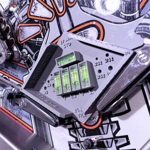 May include: A gray 3D-printed pinball machine leveling tool featuring five integrated green liquid spirit level vials. The tool is marked with text including 27 MM STD, 5 DIAMOND, and 24 N TILT, and is positioned between two metal pinball flippers.