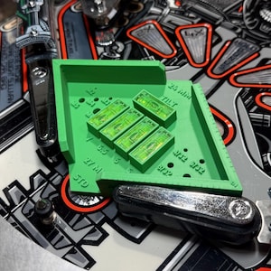 May include: A green 3D-printed pinball machine leveler with five small, clear bubble levels. The leveler sits on a pinball machine playfield, near a chrome plunger and other machine components. The leveler has measurements in inches.