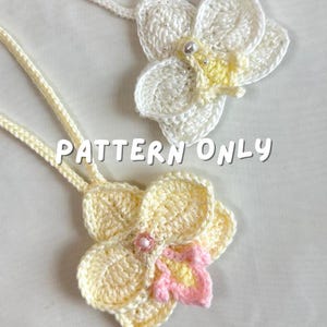 May include: Two crocheted flower pendants. One is white with yellow accents and a pearl center, the other is yellow with pink accents and a beaded center. The text "PATTERN ONLY" is visible.