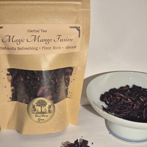 Peut inclure: Un sachet en papier brun de thé aux herbes étiqueté "Magic Mango Fusion". Le sachet est partiellement transparent, révélant les feuilles de thé. Un petit bol blanc contient une portion du thé, et un petit tas de feuilles de thé est devant.