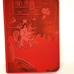 Puede incluir: Carpeta roja con una ilustración de dibujos animados de una criatura felina y un hombre con traje. La carpeta tiene la palabra "vaultx" impresa en la parte inferior. El fondo muestra una estantería y un portátil.