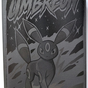 Puede incluir: Carpeta de cartas negra con Umbreon, un personaje de Pokémon, con el nombre "UMBREON" en texto estilizado. La carpeta tiene una superficie texturizada y el logotipo de Vault X en la parte inferior.