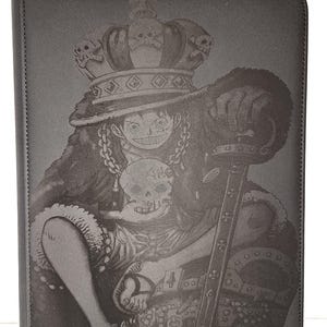 Puede incluir: Carpeta de cartas negra con una ilustración detallada de un personaje con una corona y una espada. La palabra "vaultx" está impresa en la parte inferior. La carpeta tiene una superficie texturizada y una marca de libro amarilla.