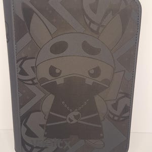 Puede incluir: Un archivador gris con un diseño de personaje de dibujos animados ninja. El archivador tiene un cierre de cremallera y un patrón repetido de la cara del personaje y otras formas. El texto "VeeTX" es visible en la parte inferior.