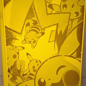 Puede incluir: Carpeta de tarjetas amarilla con Pikachu de la franquicia Pokémon. La carpeta tiene un patrón repetido de personajes Pikachu y diseños de rayos. El logotipo de Vaultix es visible en la parte inferior. La carpeta está hecha de un material texturizado.