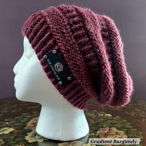 May include: A burgundy knit beanie with a textured pattern. The hat features a black label with the text "SLIP.STITCH.SNIP" and a skull graphic. The hat is displayed on a white mannequin head. The text "Gradient Burgundy" is at the bottom.