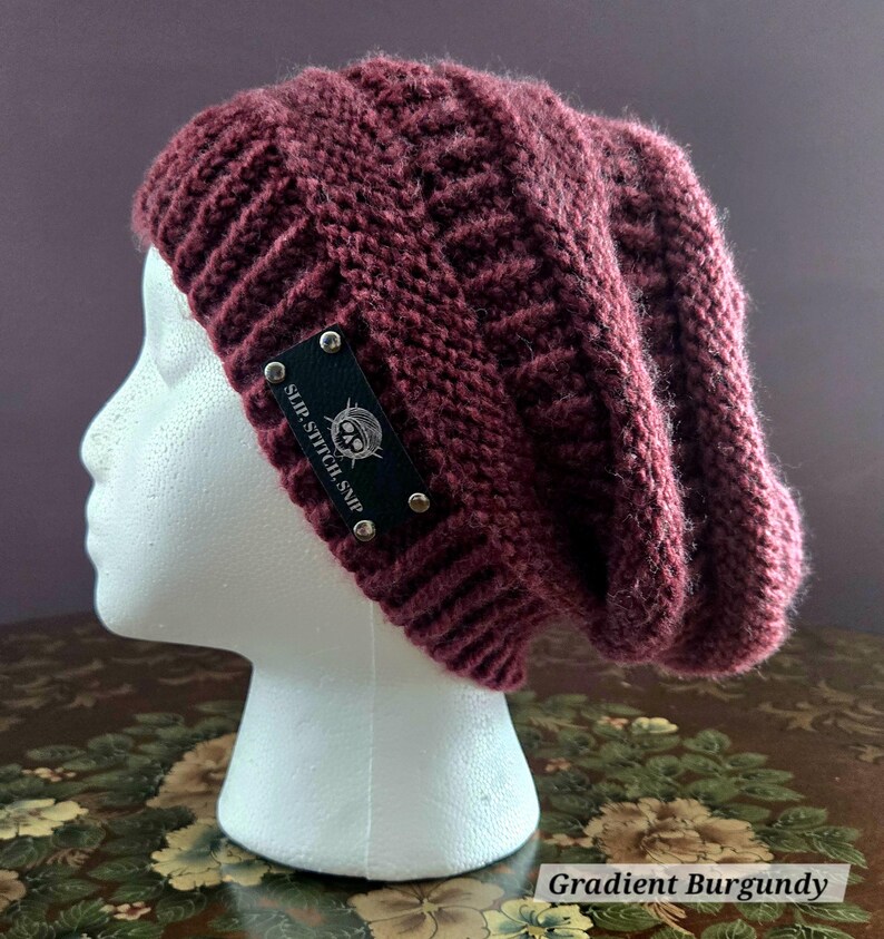 May include: A burgundy knit beanie with a textured pattern. The hat features a black label with the text "SLIP.STITCH.SNIP" and a skull graphic. The hat is displayed on a white mannequin head. The text "Gradient Burgundy" is at the bottom.