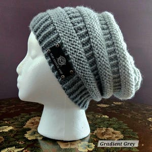 May include: A gradient grey knit beanie hat with a textured design. The hat features a black label with the text "SLIP.STITCH.SNIP" and a skull logo. The hat is displayed on a white mannequin head.