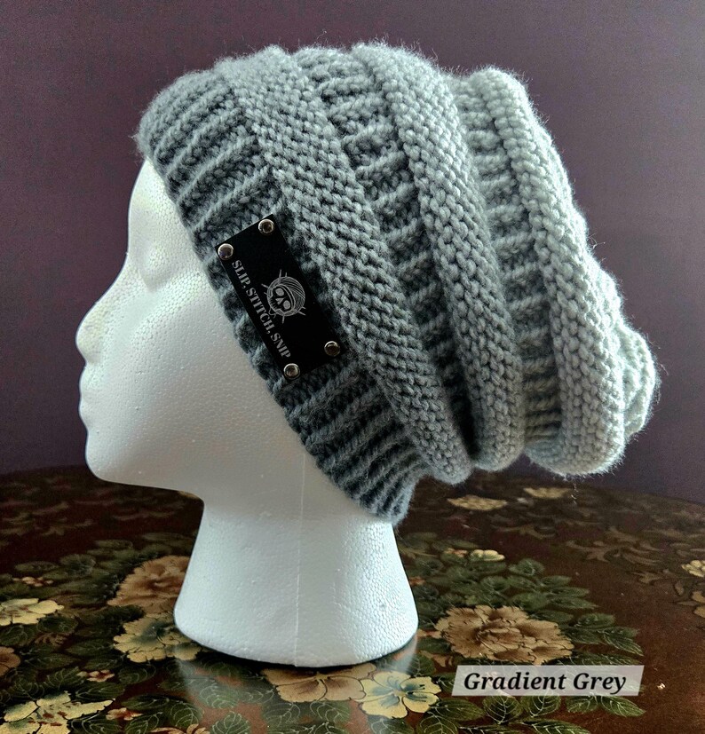 May include: A gradient grey knit beanie hat with a textured design. The hat features a black label with the text "SLIP.STITCH.SNIP" and a skull logo. The hat is displayed on a white mannequin head.