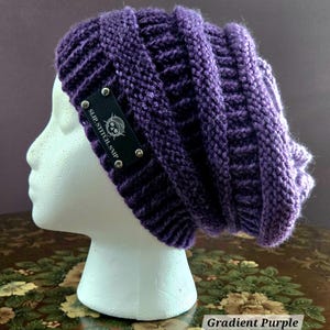 May include: A gradient purple knit beanie with a ribbed design. The beanie has a black tag with the text "SLIP.STITCH.SNIP" and a logo, secured with silver rivets. The hat is displayed on a white mannequin head.
