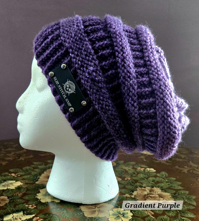 May include: A gradient purple knit beanie with a ribbed design. The beanie has a black tag with the text "SLIP.STITCH.SNIP" and a logo, secured with silver rivets. The hat is displayed on a white mannequin head.