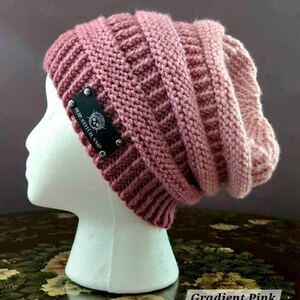 May include: A gradient pink knit beanie hat with a textured pattern. The hat features a black label with the text "SLIP. STITCH. SNIP" and a skull logo. The hat is displayed on a white mannequin head.