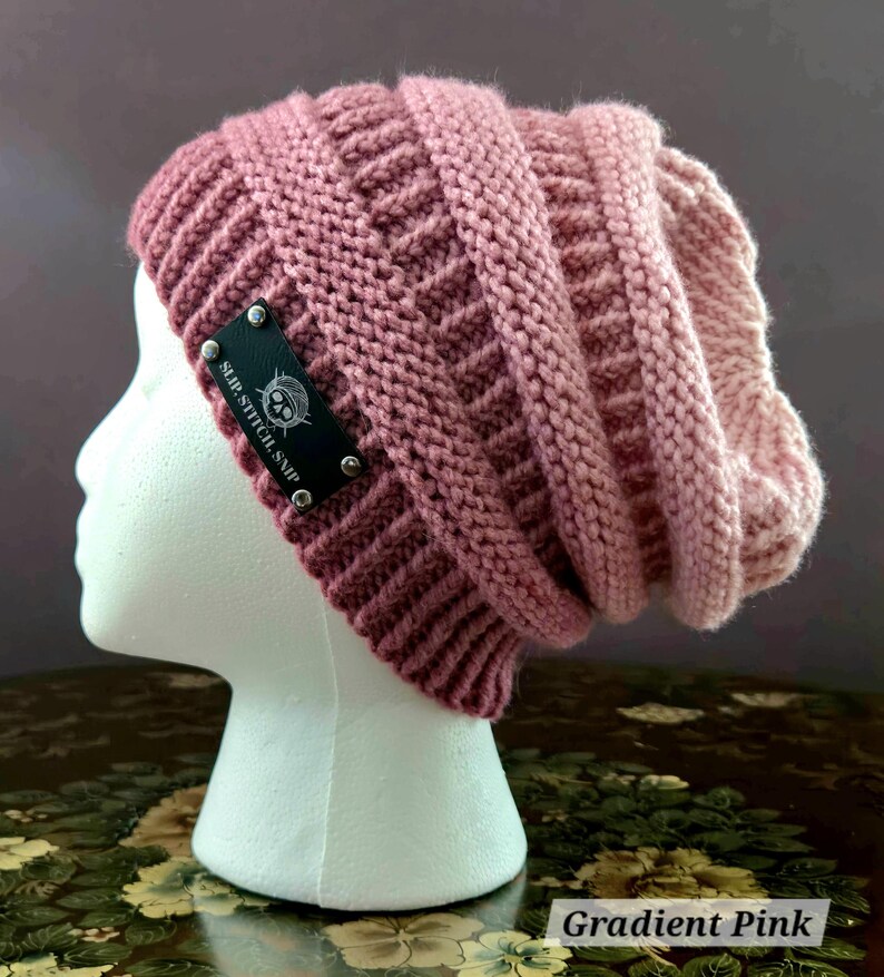 May include: A gradient pink knit beanie hat with a textured pattern. The hat features a black label with the text "SLIP. STITCH. SNIP" and a skull logo. The hat is displayed on a white mannequin head.