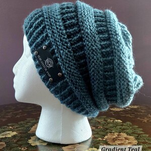 May include: A teal knitted beanie hat with a ribbed design. The hat has a small black tag with silver rivets and the text "SLIP.STITCH.SNIP". The hat is displayed on a white mannequin head.