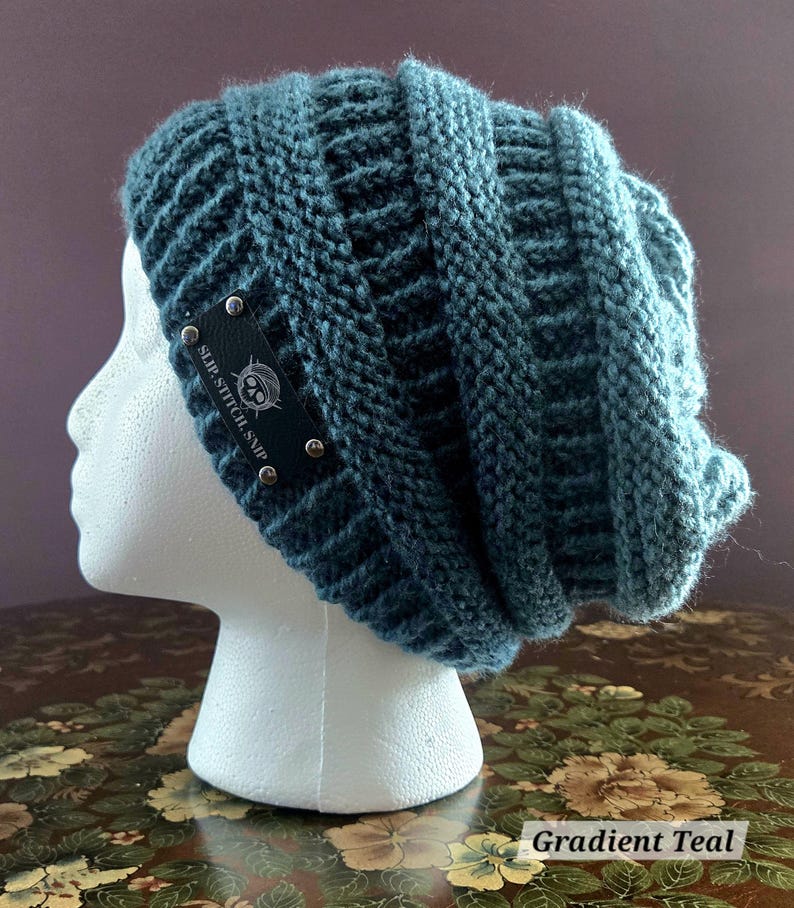May include: A teal knitted beanie hat with a ribbed design. The hat has a small black tag with silver rivets and the text "SLIP.STITCH.SNIP". The hat is displayed on a white mannequin head.
