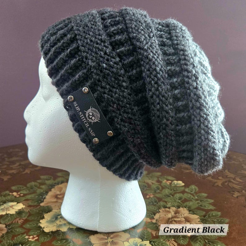 May include: A dark gray knit beanie with a gradient effect. The hat features a ribbed design and a small black tag with the text "Slip Stitch SMP" and a skull logo. The hat is displayed on a white mannequin head.