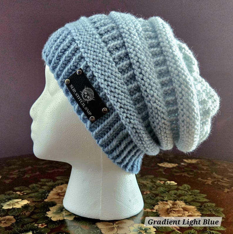 May include: A light blue knit beanie with a gradient effect. The hat features a textured pattern and a black label with the text "SLIP STITCH, SMP" and a skull logo. The text "Gradient Light Blue" is also visible.